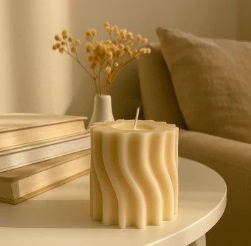 Wavy-textured candle on a table with books and a vase of dried flowers in the background.